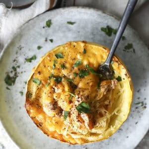 A baked Buffalo chicken spaghetti squash half filled with a cheesy, golden topping and garnished with chopped parsley sits on a plate, with a fork twirling the squash strands.