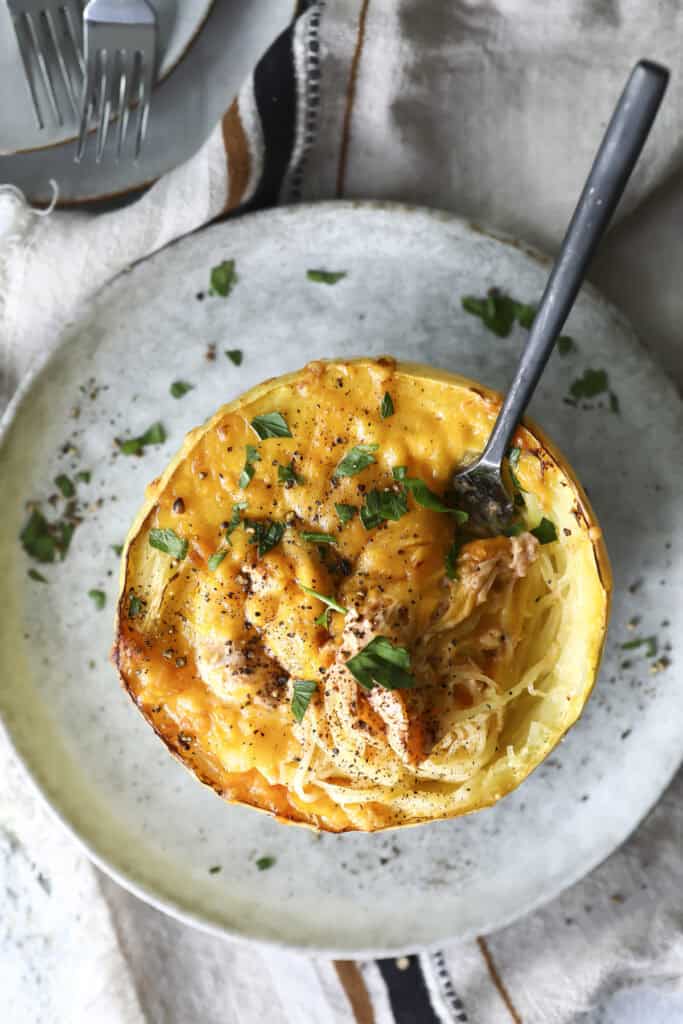 A baked Buffalo chicken spaghetti squash half filled with a cheesy, golden topping and garnished with chopped parsley sits on a plate, with a fork twirling the squash strands.