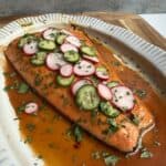 A Chipotle honey salmon fillet on a white platter, topped with sliced radishes, cucumbers, jalapeños, and fresh herbs, served in a reddish-brown sauce.