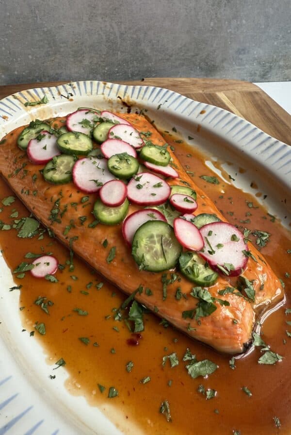 A Chipotle honey salmon fillet on a white platter, topped with sliced radishes, cucumbers, jalapeños, and fresh herbs, served in a reddish-brown sauce.