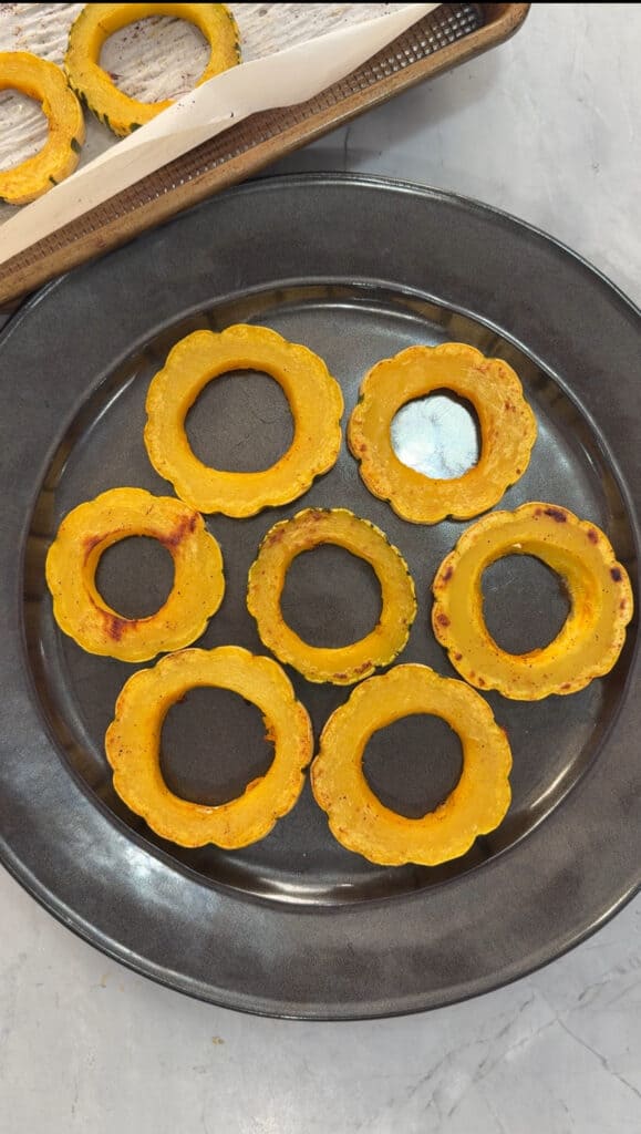 Seven roasted delicata squash rings are arranged on a dark grey plate, with a baking tray of additional squash slices visible in the background on a light-colored countertop.