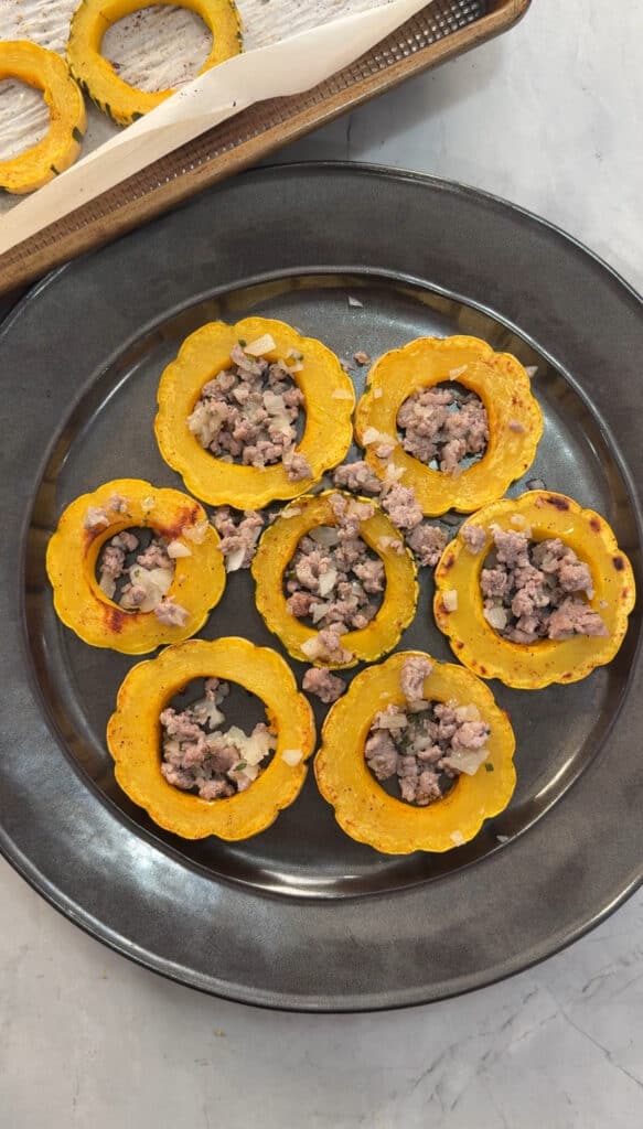 Seven roasted yellow squash rings stuffed with ground meat and chopped onions are arranged on a dark gray plate, set on a light-colored surface.