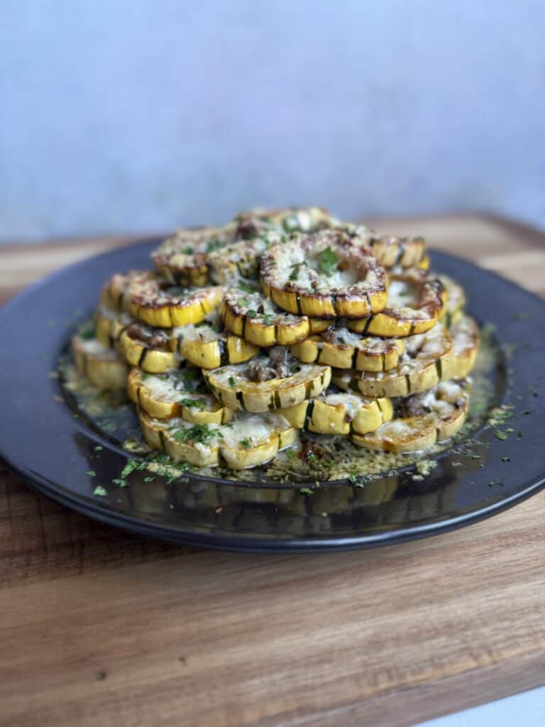 A baked delicata squash stack garnished with herbs and grated cheese is arranged on a black plate, set on a wooden surface with a neutral background.