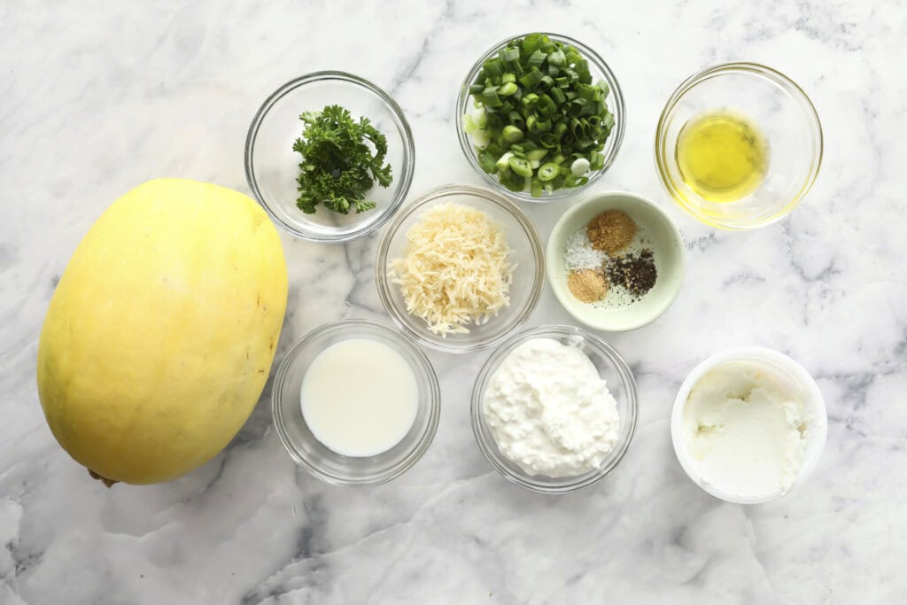 Several ingredients in glass bowls on a marble surface, including chopped parsley, sliced green onions, olive oil, shredded cheese, seasonings, milk, cottage cheese, ricotta cheese, and a whole spaghetti squash.
