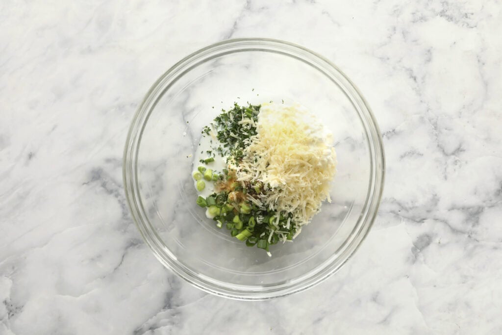 A glass mixing bowl on a marble countertop contains chopped green onions, shredded cheese, chopped herbs, and a creamy white ingredient.