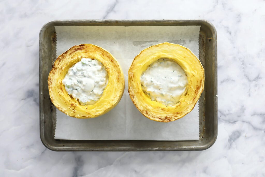 Two roasted spaghetti squash halves filled with a creamy white sauce, placed side by side on a parchment-lined metal baking tray on a marble surface.