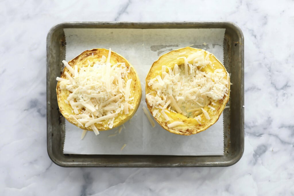 Two halves of spaghetti squash, roasted and topped with shredded cheese, sit on a parchment-lined metal baking tray on a marble countertop.