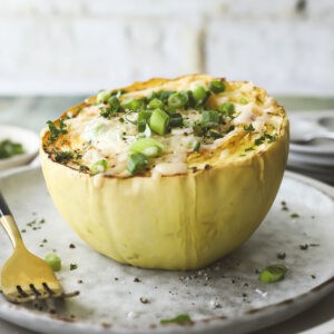 A baked French onion soup spaghetti squash half filled with melted cheese and topped with sliced green onions sits on a plate, with a fork beside it and some herbs sprinkled around.