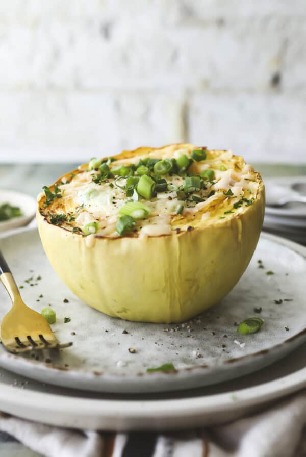 A baked French onion soup spaghetti squash half filled with melted cheese and topped with sliced green onions sits on a plate, with a fork beside it and some herbs sprinkled around.