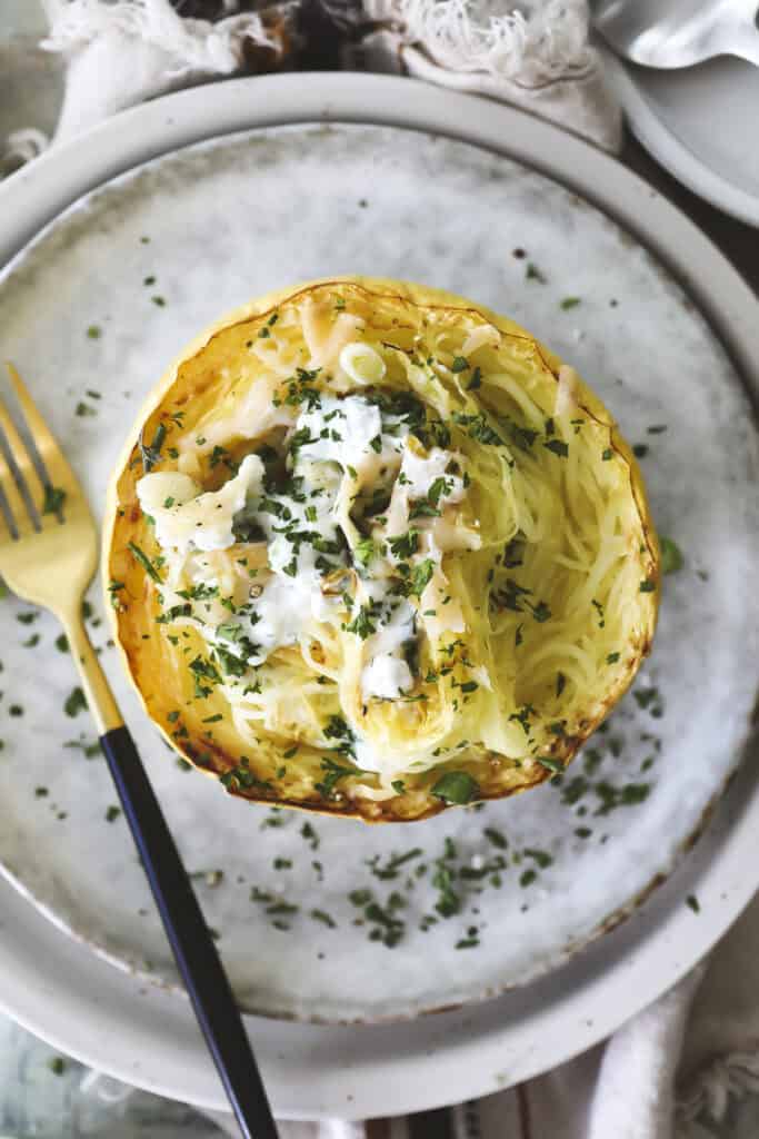 A baked French Onion Soup spaghetti squash half is served on a plate, filled with shredded squash, creamy white sauce, herbs, and garnished with chopped parsley. A gold and black fork sits beside the squash on the plate.