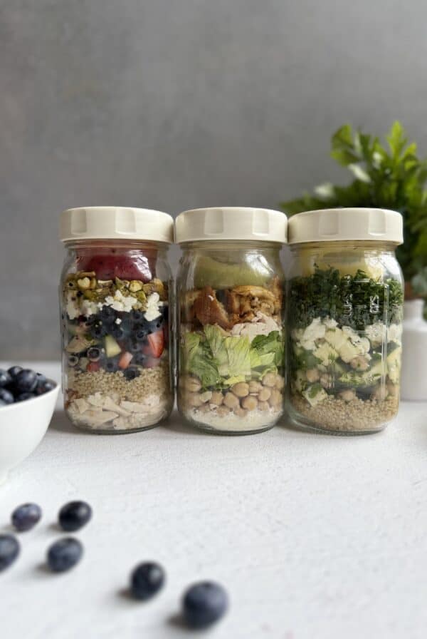 3 reset salads in glass jars with white lids filled with layered salads, each featuring grains, vegetables, and toppings. A bowl and scattered blueberries are in the foreground, with a green plant blurred in the background.