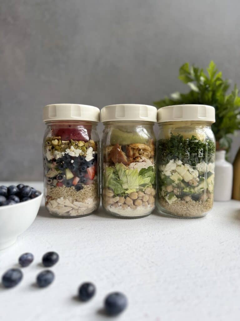 3 reset salads in glass jars with white lids filled with layered salads, each featuring grains, vegetables, and toppings. A bowl and scattered blueberries are in the foreground, with a green plant blurred in the background.