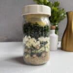 A glass jar filled with layered salad ingredients, including couscous, chickpeas, zucchini, cheese, kale, and a lemon wedge, sits on a white surface with herbs and bottles blurred in the background.