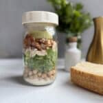 A glass jar is filled with layered salad ingredients, including chickpeas, spinach, chicken, bacon, avocado, and dressing. The jar sits on a white surface near a block of Parmesan cheese and green herbs in the background.