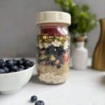 A glass jar filled with layers of oats, strawberries, blueberries, yogurt, and nuts sits on a white surface. A small bowl of blueberries and a green leafy plant are in the background.