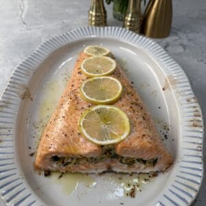 A crab stuffed salmon tail topped with four lemon slices, garnished with herbs, is served on a white oval platter with blue stripes. Two gold pepper mills and fresh herbs are in the background.