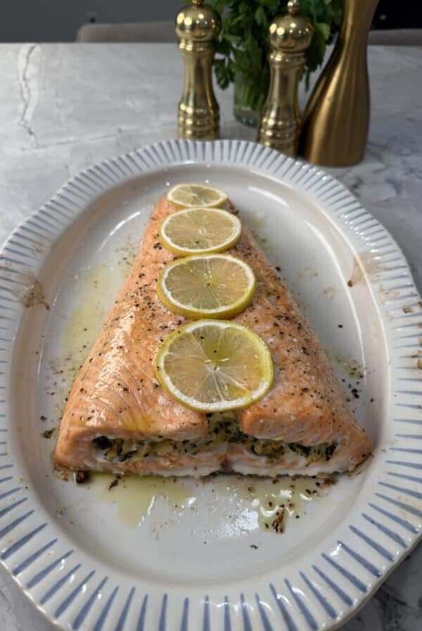 A crab stuffed salmon tail topped with four lemon slices, garnished with herbs, is served on a white oval platter with blue stripes. Two gold pepper mills and fresh herbs are in the background.