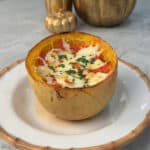 A halved spaghetti squash filled with melted cheese, herbs, and sauce sits on a white plate with a brown rim. Decorative gold pumpkins are in the background on a marble surface.