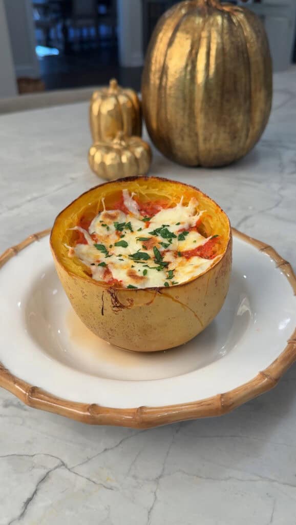 A halved spaghetti squash filled with melted cheese, herbs, and sauce sits on a white plate with a brown rim. Decorative gold pumpkins are in the background on a marble surface.