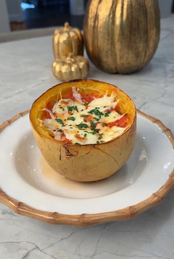 A halved spaghetti squash filled with melted cheese, herbs, and sauce sits on a white plate with a brown rim. Decorative gold pumpkins are in the background on a marble surface.