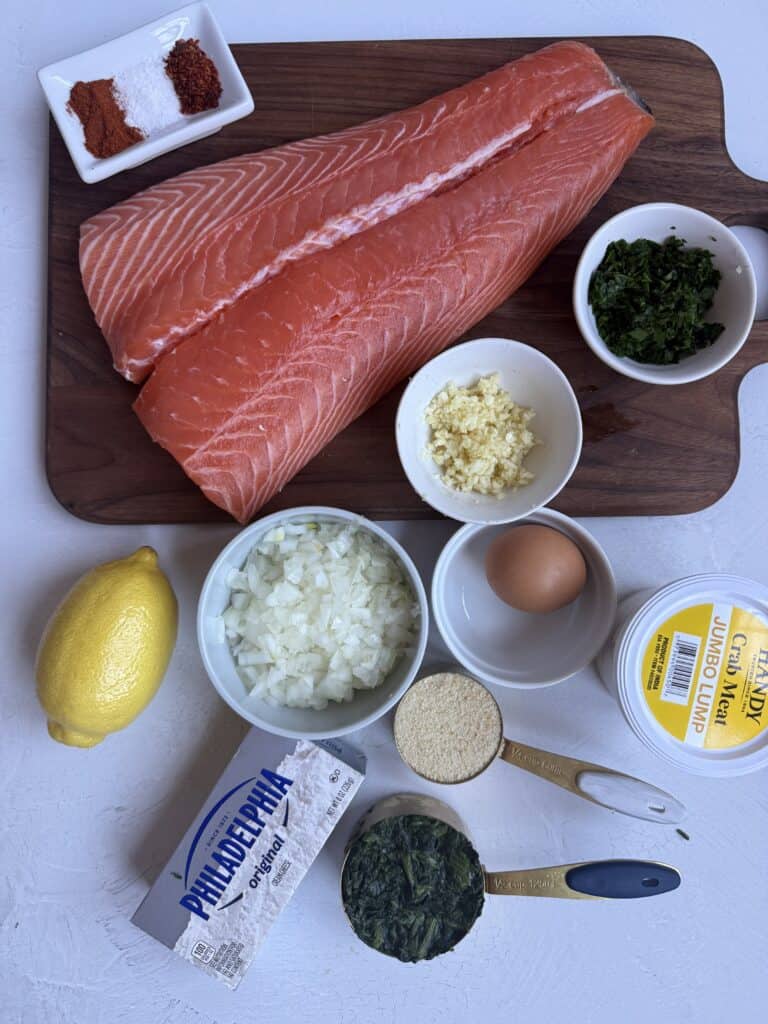 Ingredients for crab stuffed salmon tail dish arranged on a white surface, including raw salmon fillets, chopped onion, minced garlic, egg, parsley, lemon, spices, panko breadcrumbs, cream cheese, butter, and frozen spinach.