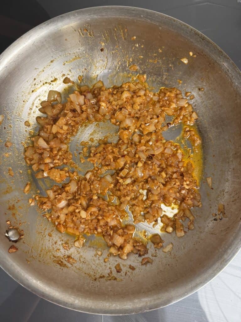 Chopped onions and garlic sautéed with spices in a stainless steel frying pan, with a golden-brown color and traces of oil and seasoning visible.