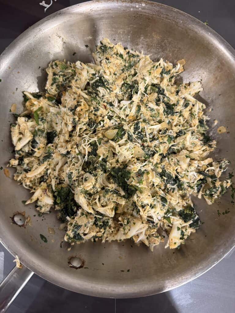 A large metal skillet filled with a mixture of shredded cooked crab, chopped leafy greens, and herbs, all lightly sautéed and mixed together.