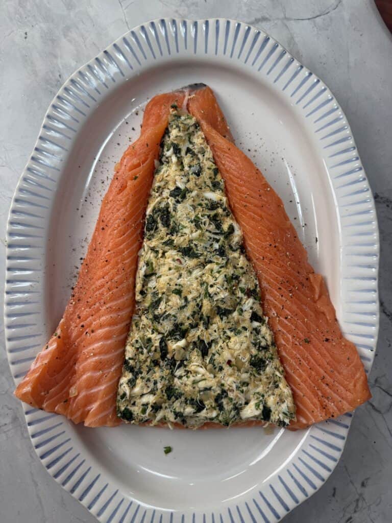 A fillet of raw salmon is partially folded open on a white oval plate, revealing a crab, spinach and cream cheese filling inside. The dish is sprinkled with black pepper and placed on a marble surface.
