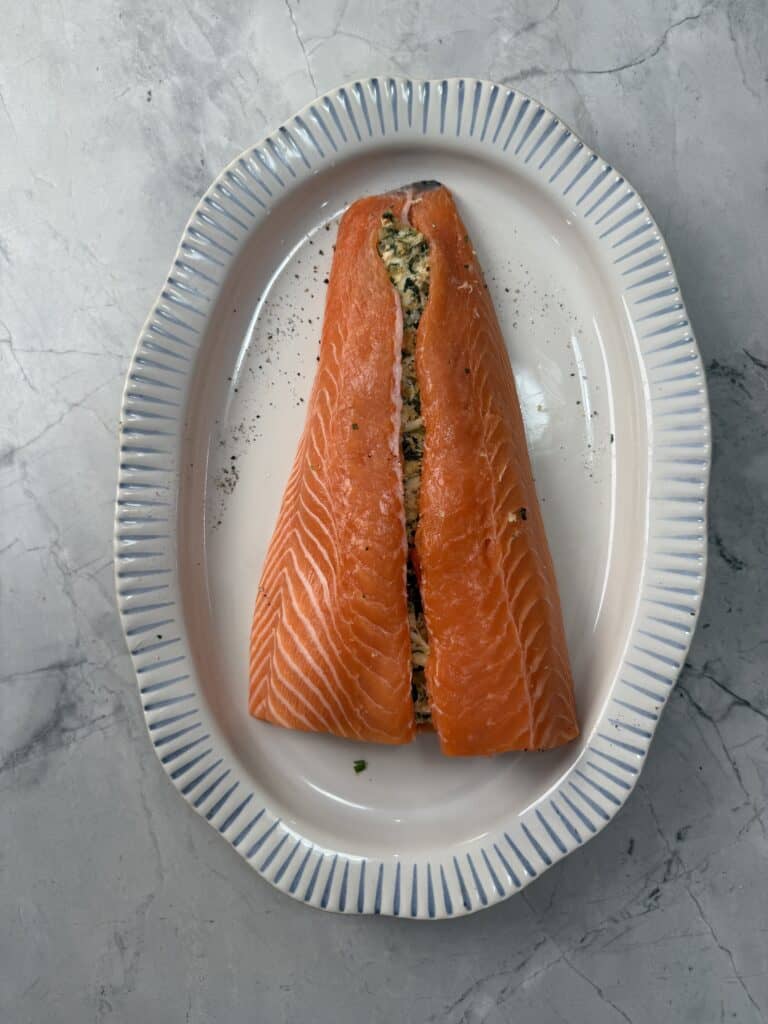 A white oval dish with blue trim holds a raw salmon fillet sliced open and stuffed with a creamy herb filling. The dish sits on a gray marbled surface.