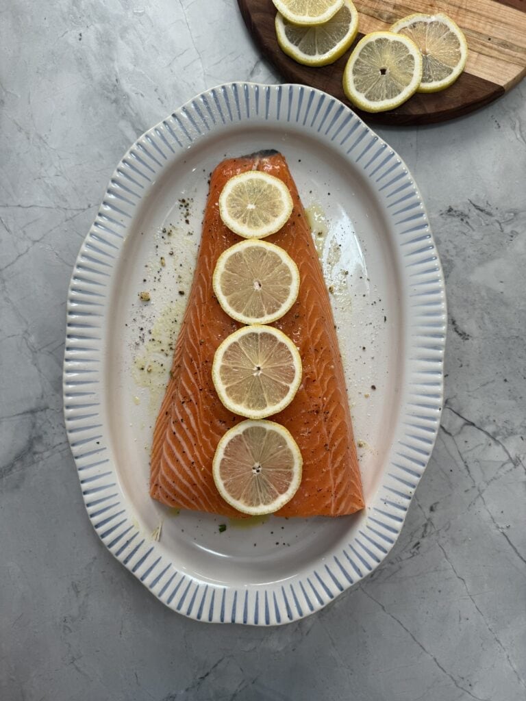 A raw salmon fillet topped with five lemon slices is arranged on a white oval dish with blue stripes. In the background, more lemon slices rest on a wooden board. The surface is gray marble.