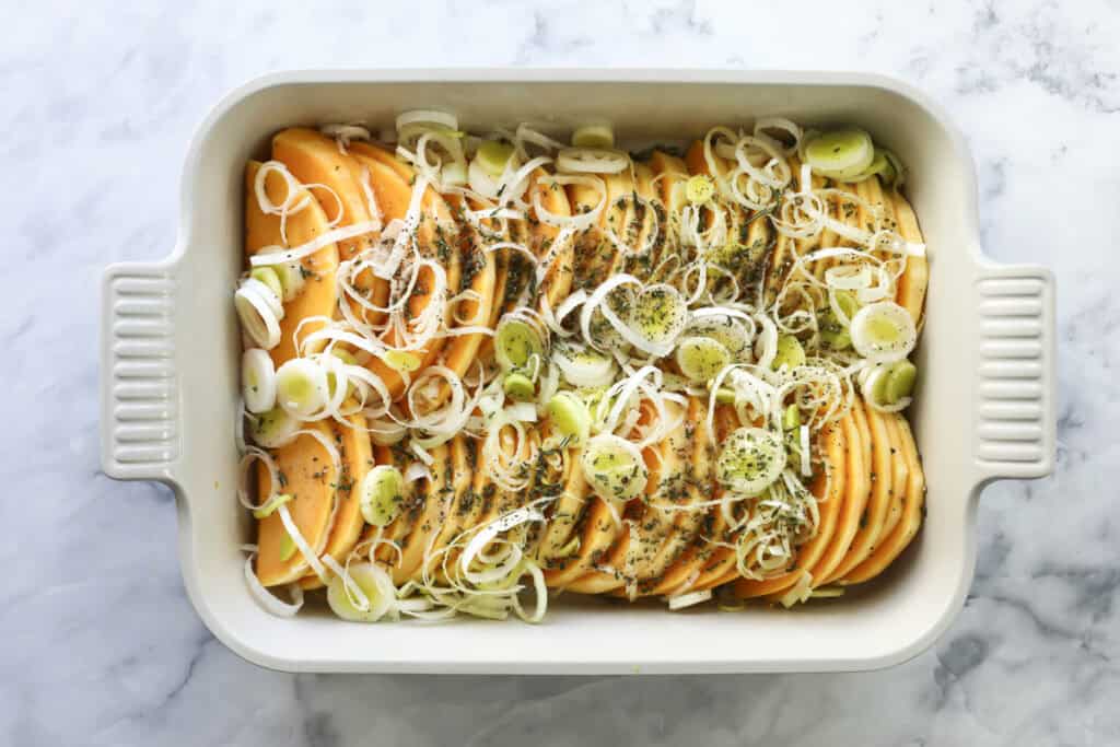 A white baking dish filled with thinly sliced butternut squash arranged in rows, topped with sliced leeks and sprinkled with herbs, ready to be baked. The dish sits on a marble countertop.