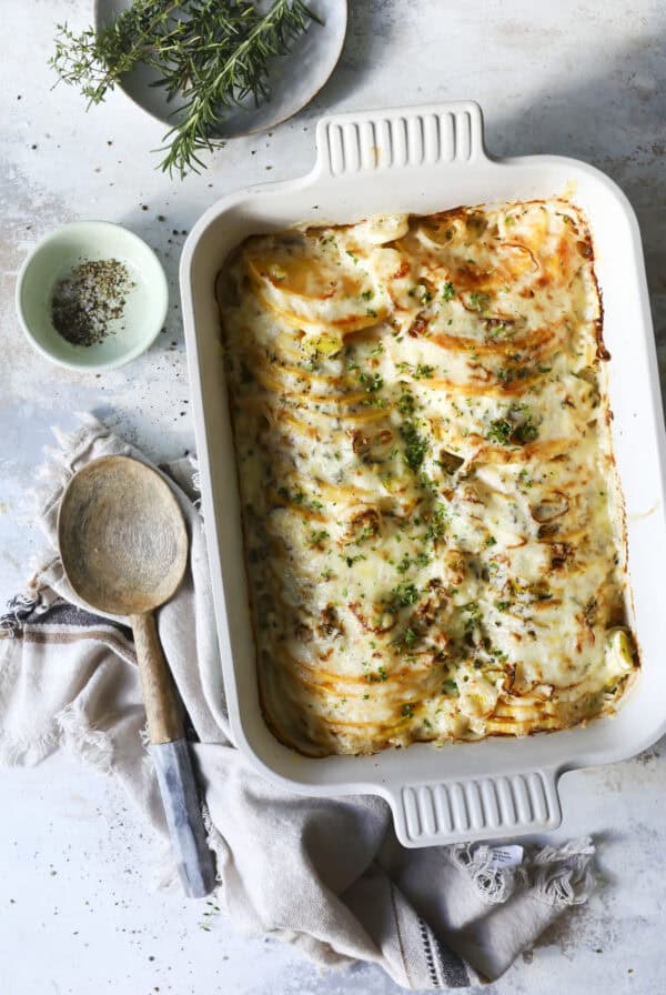 An easy Butternut Squash Gratin baked in a casserole dish, topped with melted cheese and chopped herbs sits in a white ceramic dish. A wooden spoon, cloth napkin, and small bowls of spices and fresh herbs are arranged nearby on a textured surface.