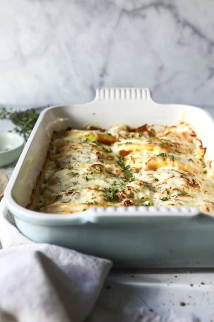 A easy butternut squash gratin baked in a casserole dish filled with cheesy, golden-brown lasagna topped with melted cheese and fresh herbs, sitting on a light-colored countertop with a napkin nearby.