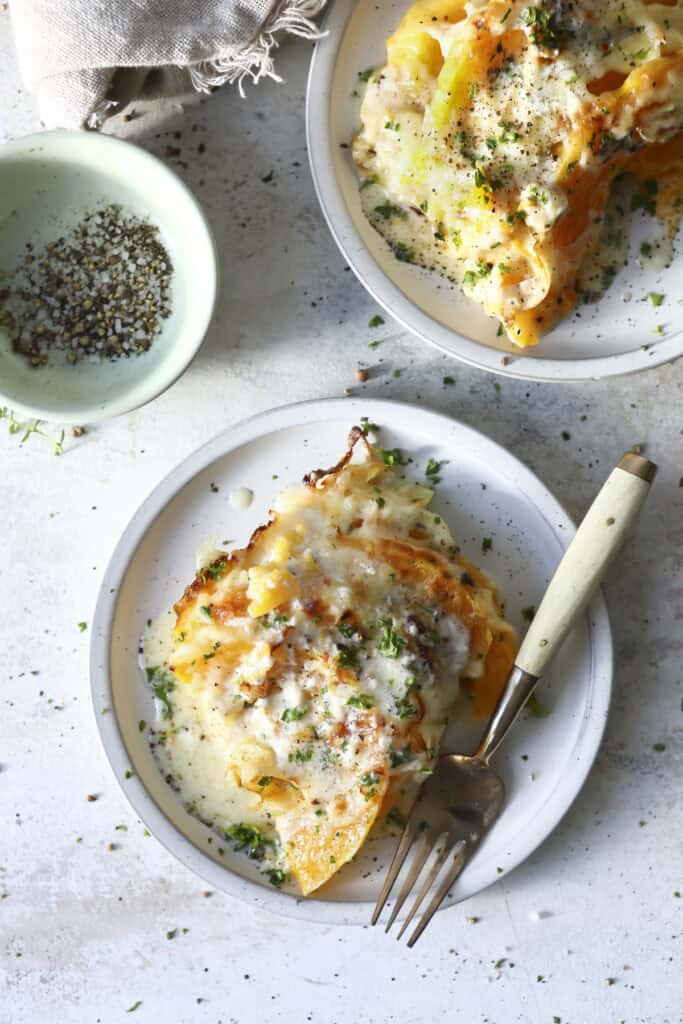 Two plates of easy butternut squash gratin, baked, topped with herbs and creamy white sauce, with a fork on one plate. A small dish of ground pepper and a cloth napkin are nearby on a light-colored surface.