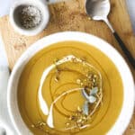 A bowl of creamy yellow Hot Honey Butternut Squash soup garnished with a swirl of cream, chopped pistachios, and fresh herbs sits on a wooden board with a spoon nearby and a small dish of salt and pepper. Toasted bread is partially visible in the corner.
