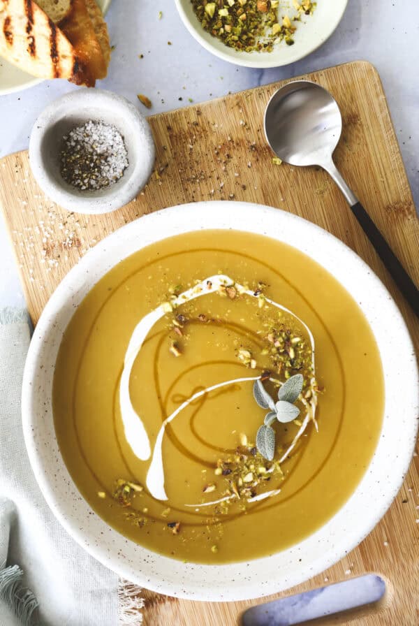 A bowl of creamy yellow Hot Honey Butternut Squash soup garnished with a swirl of cream, chopped pistachios, and fresh herbs sits on a wooden board with a spoon nearby and a small dish of salt and pepper. Toasted bread is partially visible in the corner.