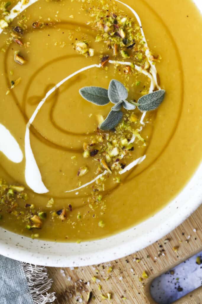 A close-up of a bowl of creamy orange hot honey butternut squash soup garnished with a swirl of cream, chopped pistachios, and fresh sage leaves, set on a wooden surface with a blue napkin partially visible.