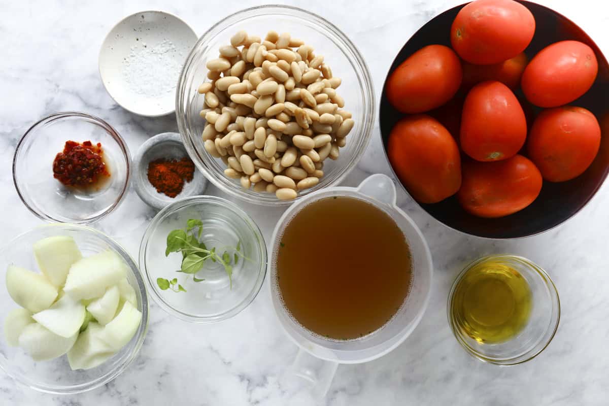 A top view of various ingredients on a marble surface, including white beans, roma tomatoes, chopped onion, vegetable broth, olive oil, fresh herbs, chili paste, paprika, and salt.