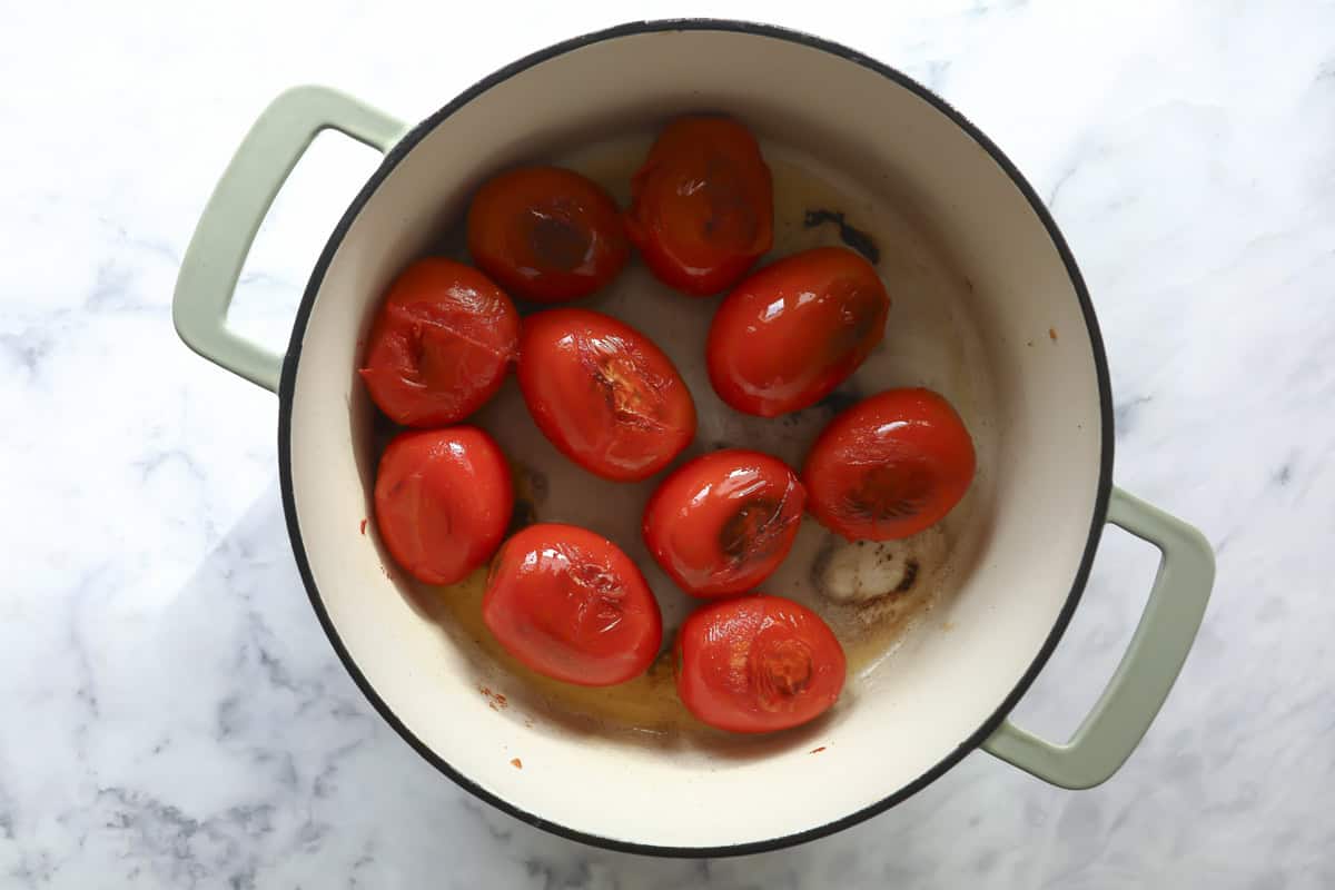 A green-handled pot containing peeled, roasted tomatoes sits on a white marble surface. The tomatoes are lightly browned and arranged in a single layer inside the pot.