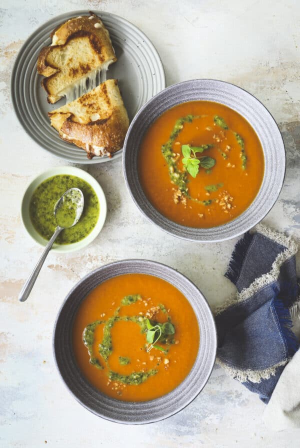 Two bowls of tomato soup garnished with green herb oil and fresh herbs, served with a plate of grilled cheese sandwiches and a small bowl of extra herb oil with a spoon, on a light textured surface.