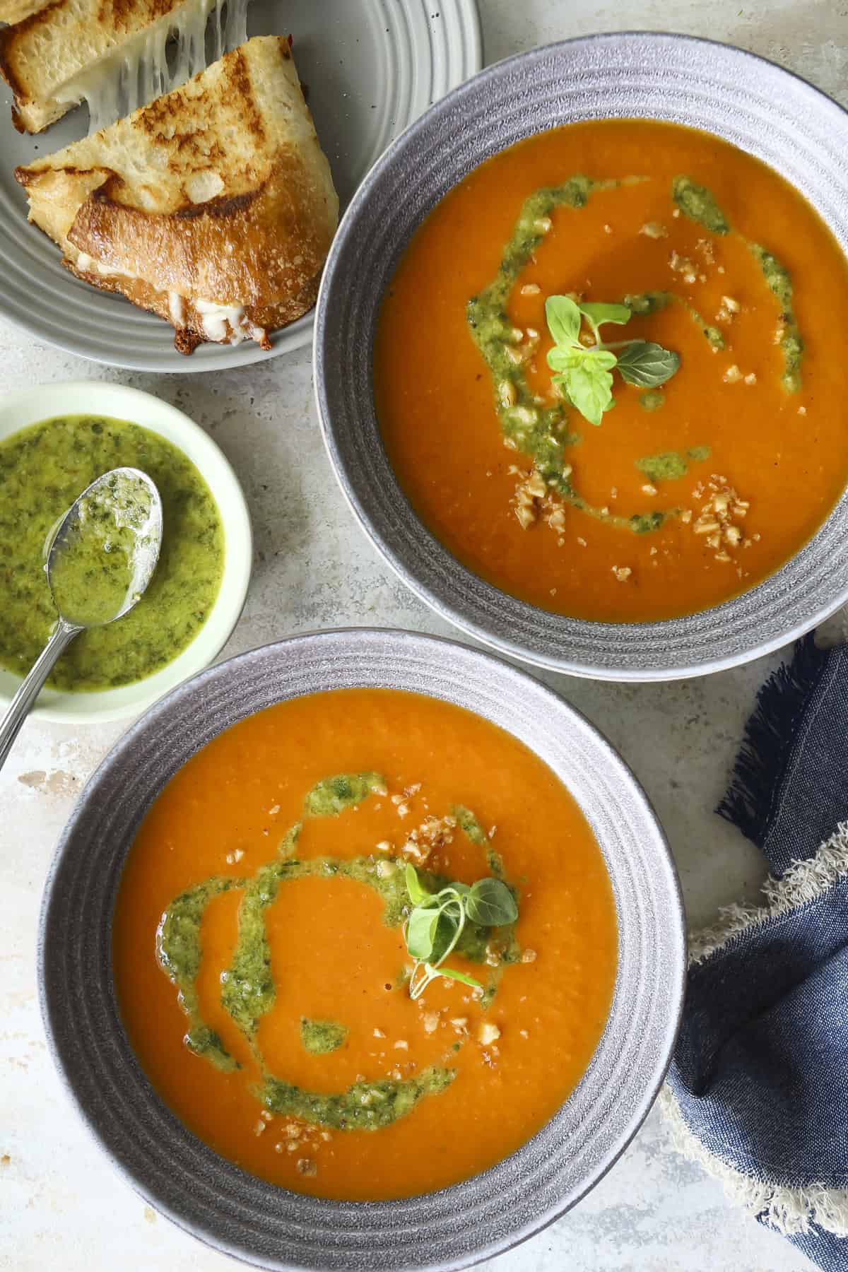 Two bowls of Charred Tomato and Walnut Pesto Soup garnished with green pesto and fresh basil sit on a table, accompanied by a plate of grilled cheese sandwiches and a small bowl of pesto with a spoon.