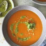 A bowl of vibrant orange tomato soup topped with a swirl of green pesto and microgreens, placed on a light-colored surface with a small dish of extra pesto nearby.