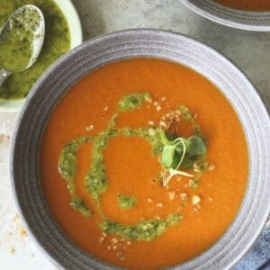 A bowl of vibrant orange tomato soup topped with a swirl of green pesto and microgreens, placed on a light-colored surface with a small dish of extra pesto nearby.