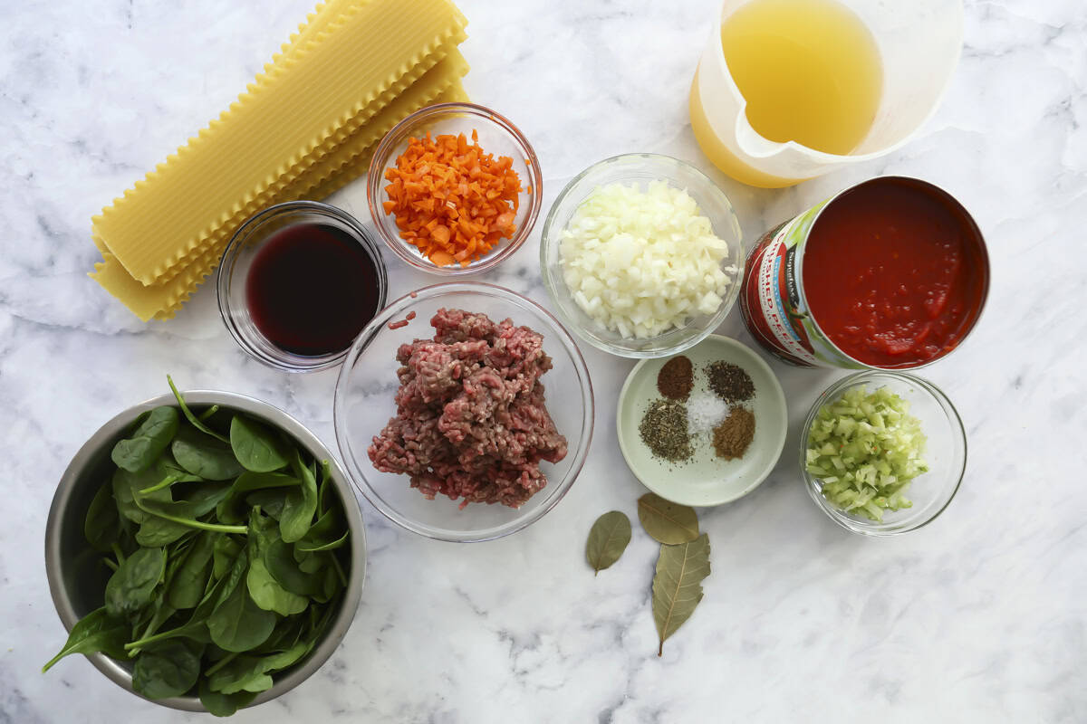 Various ingredients for a pasta dish are arranged on a marble surface, including lasagna noodles, ground meat, spinach, chopped onion, chopped carrot, celery, tomato sauce, red wine, herbs, and a jug of broth.