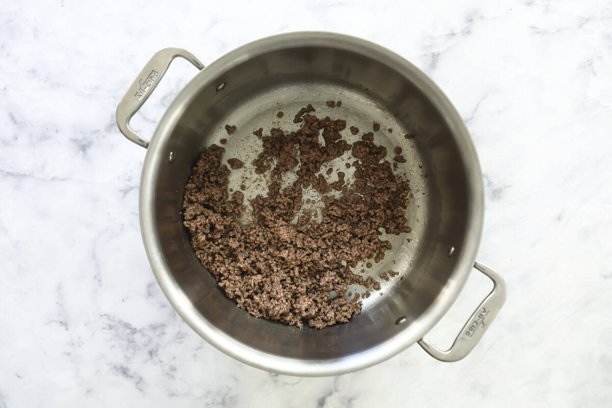 Ground beef is being browned in a large stainless steel pot on a white marble countertop. The beef is partially cooked and spread out in the center and sides of the pot.