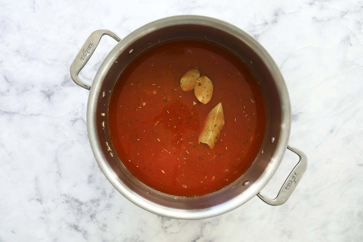 A stainless steel pot filled with red tomato sauce, bay leaves, and a halved garlic bulb, placed on a white marble surface.