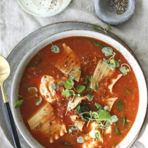 A bowl of Lasagna Bolognese soup with lasagna noodles, spinach, and fresh herbs, topped with cracked black pepper. Beside it are bowls of grated cheese, sour cream, and more pepper on a linen surface.