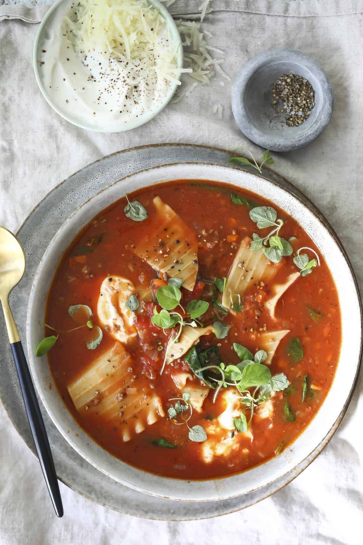 A bowl of Lasagna Bolognese soup with lasagna noodles, spinach, and fresh herbs, topped with cracked black pepper. Beside it are bowls of grated cheese, sour cream, and more pepper on a linen surface.
