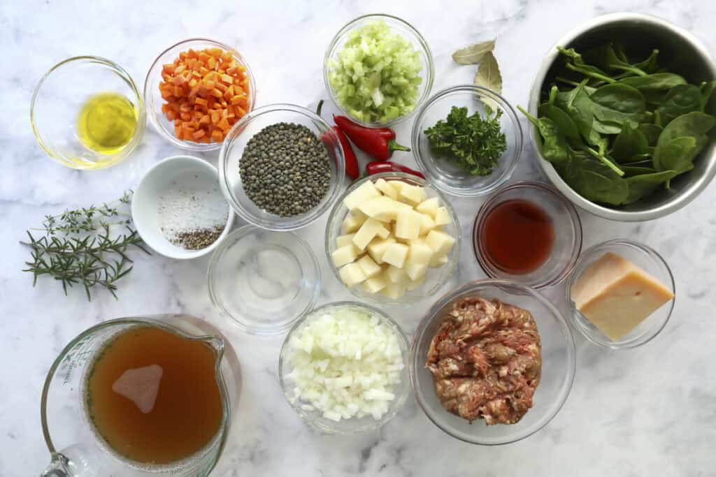 A top-down view of various ingredients in glass bowls on a marble surface, including chopped vegetables, raw sausage, lentils, fresh spinach, cheese, herbs, broth, and seasonings.