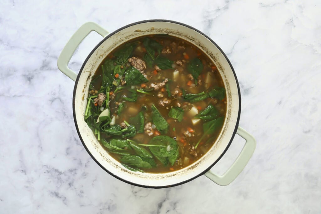 A green-handled pot filled with soup containing ground meat, spinach leaves, diced carrots, and potatoes sits on a light marble surface.